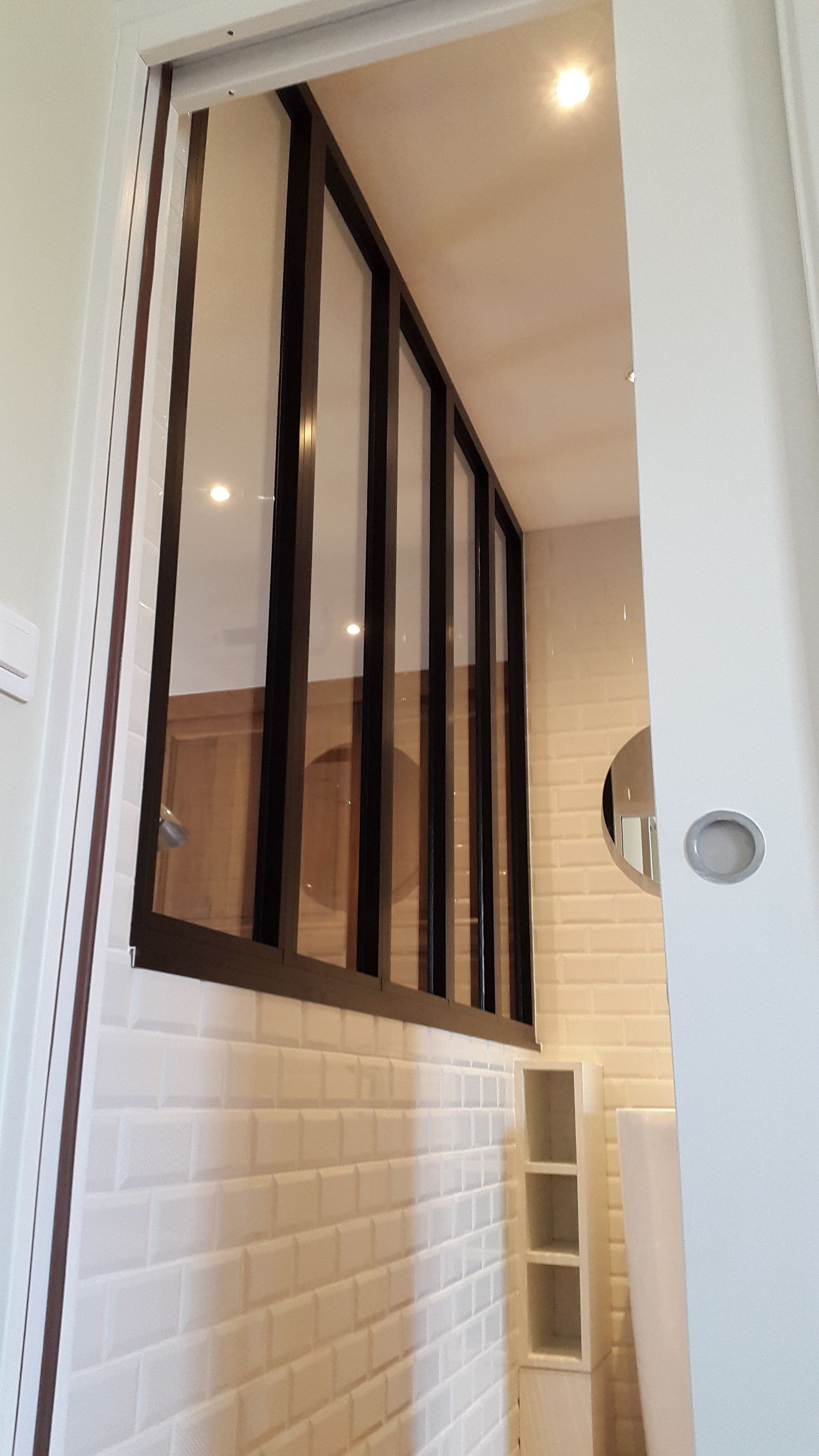 Vue intérieure à travers un cadre de porte blanc ouvert, montrant une cloison vitrée avec des cadres en métal foncé au-dessus de carreaux de métro blancs.