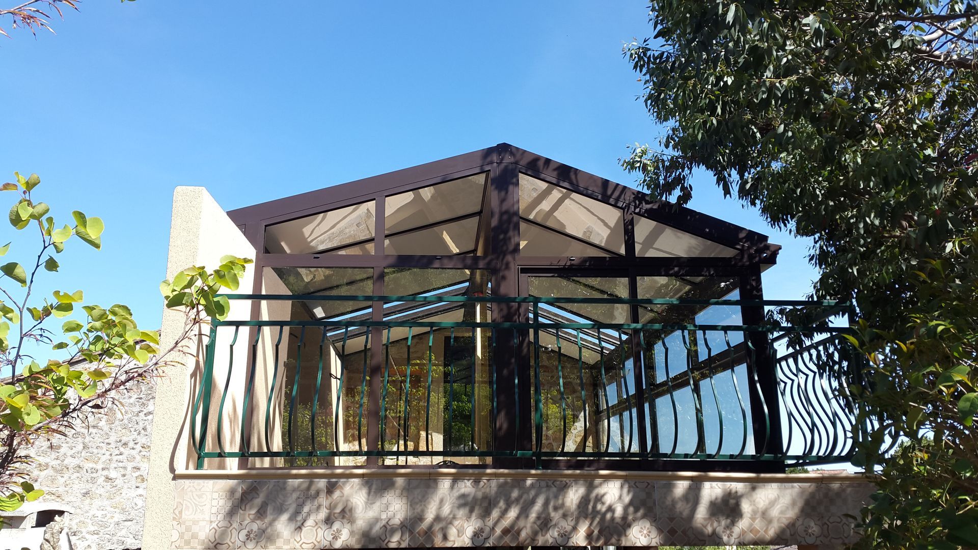 Une terrasse surélevée et couverte, avec une structure sombre, des parois vitrées et une balustrade décorative verte, se détachant sur un ciel d'un bleu éclatant.