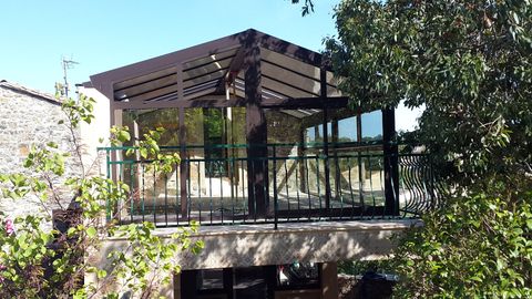 Un bâtiment en pierre avec une terrasse agrémentée d'une pergola en bois et d'une balustrade en métal sombre, entourée d'une végétation luxuriante.