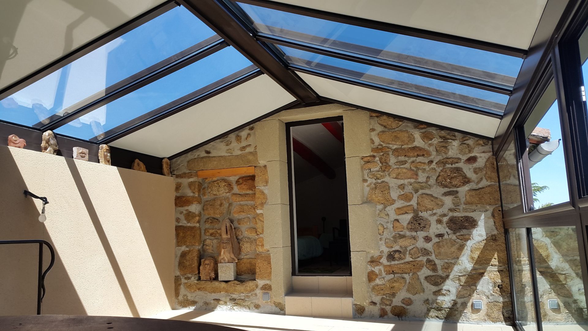 Vue intérieure d'une véranda présentant un mur en pierre, une porte centrale et un toit en verre incliné.