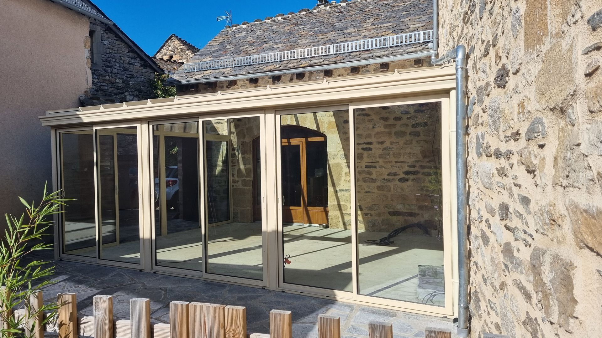Une véranda avec des cadres beiges et des portes coulissantes en verre, attenante à un bâtiment en pierre.
