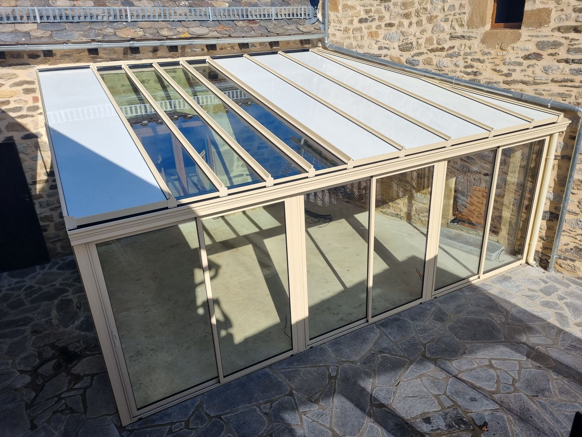 Une véranda beige rétractable, aux parois vitrées et au toit partiellement ouvert, est installée sur une terrasse en pierre, adossée à un mur en pierre.