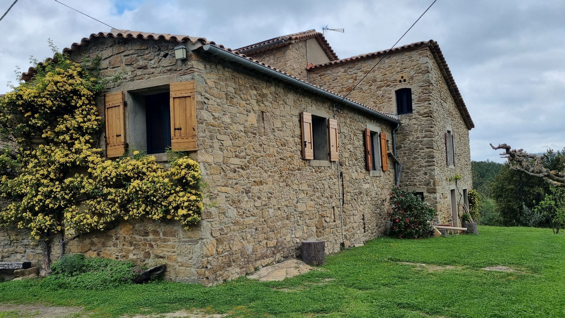 Une ferme rustique en pierre, avec des volets en bois et une végétation grimpante, située dans un paysage verdoyant et herbeux.