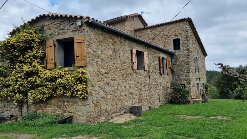 Une maison rustique en pierre à plusieurs niveaux, avec des volets en bois et une plante grimpante jaune-vert, située dans un paysage herbeux.