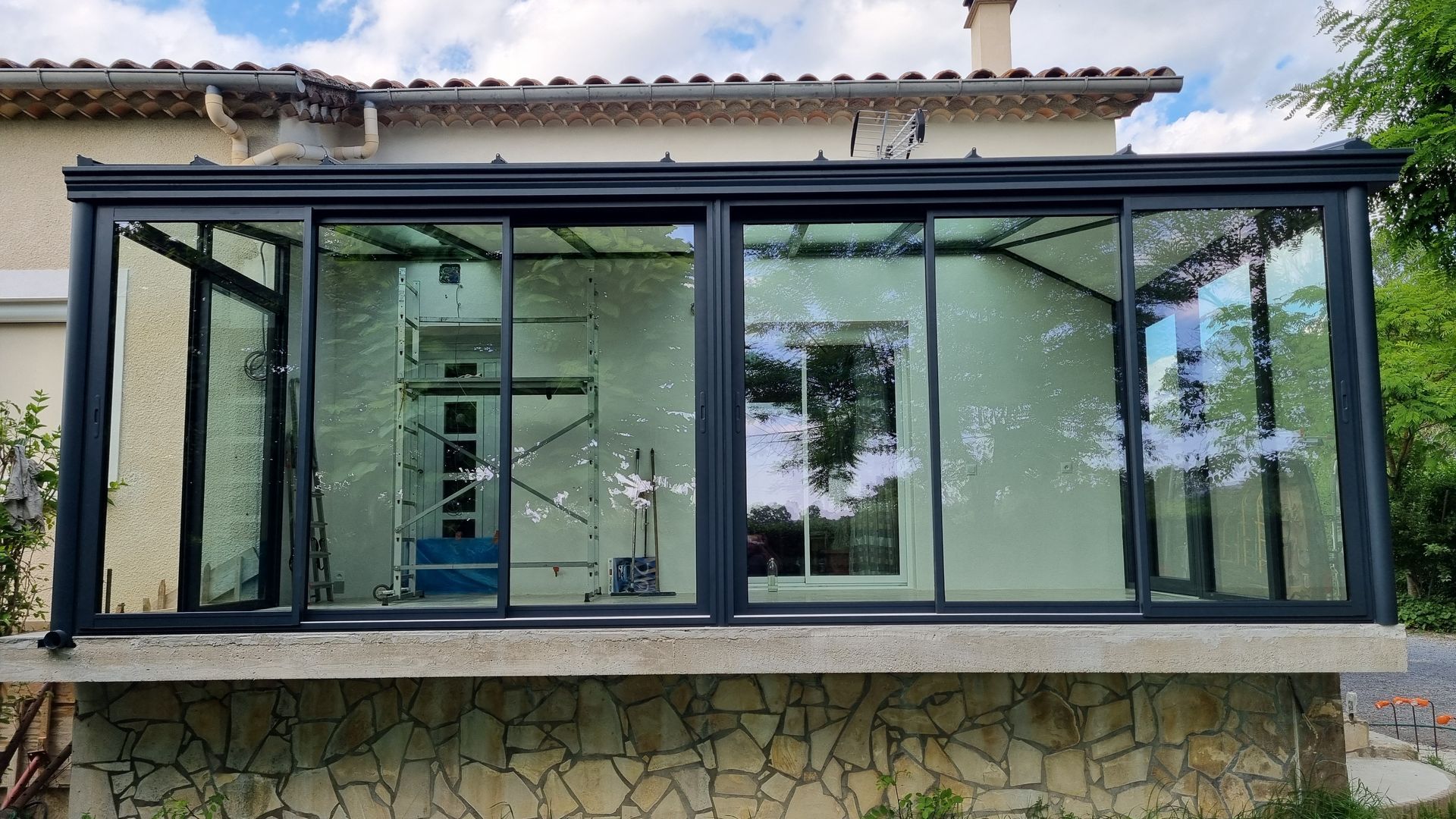 Une véranda en verre à la structure sombre, adossée à une maison à soubassement en pierre et dont l'intérieur laisse apparaître des échafaudages.