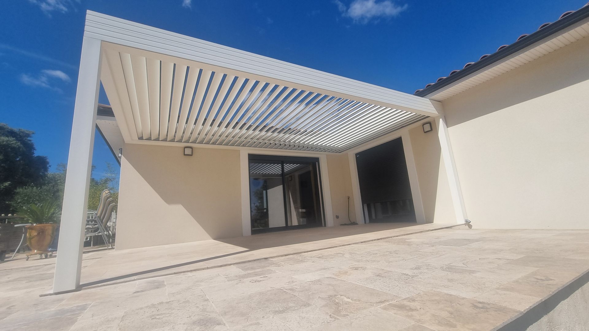 Une pergola bioclimatique blanche à persiennes réglables, fixée à une maison au-dessus d'une terrasse en pierre, sous un ciel bleu azur.
