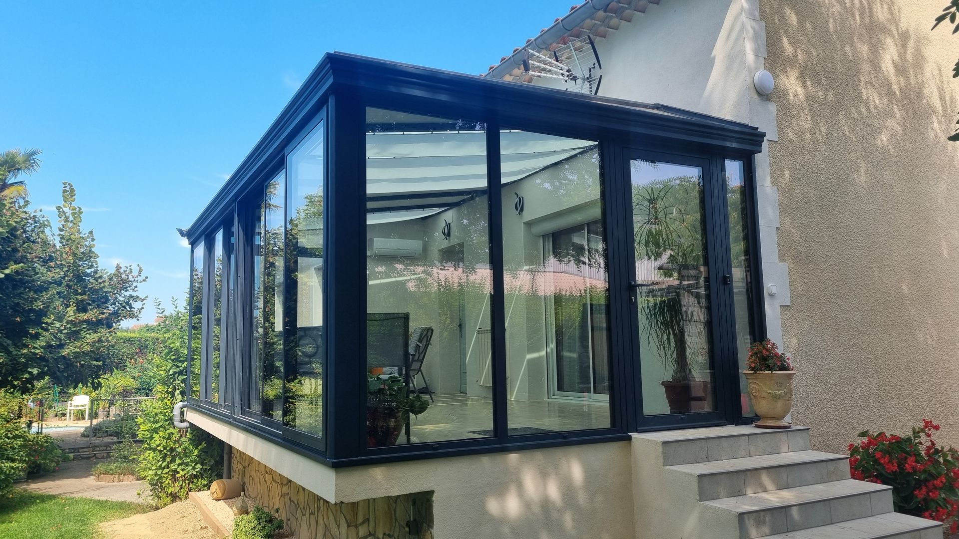 Une véranda moderne en verre à cadre noir, attenante à une maison avec des marches en béton menant à l'entrée latérale.