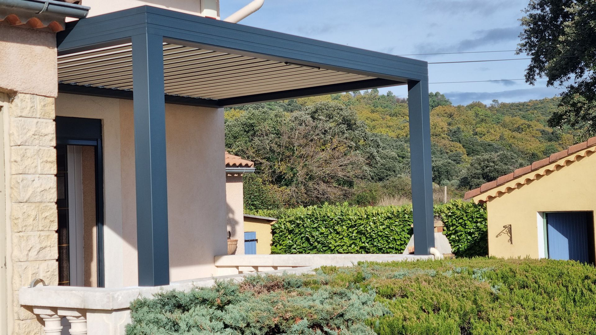 Une pergola moderne gris foncé avec un auvent rétractable, adossée à une maison en pierre donnant sur un jardin.