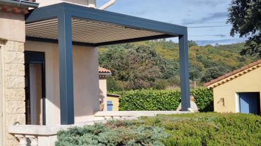 Une pergola en métal gris foncé, dotée d'une toile rétractable, s'étend d'une maison en stuc clair vers un jardin.