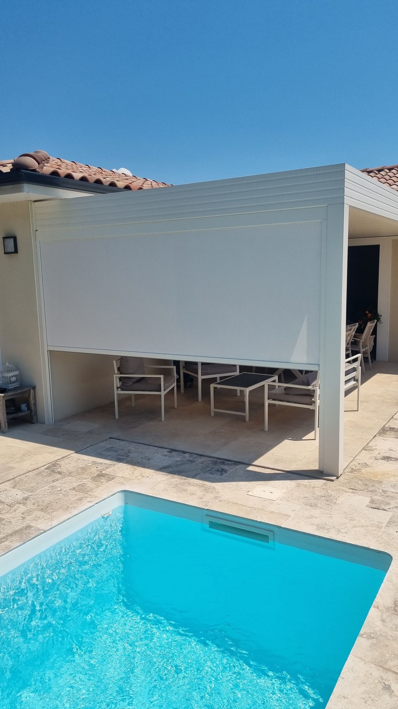 Un store rétractable blanc recouvre le côté d'un coin salon extérieur jouxtant une piscine d'un bleu éclatant.