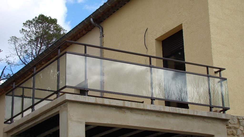 Un immeuble beige avec un balcon doté d'une rambarde en métal noir et de panneaux de verre transparent, sous un ciel bleu nuageux.