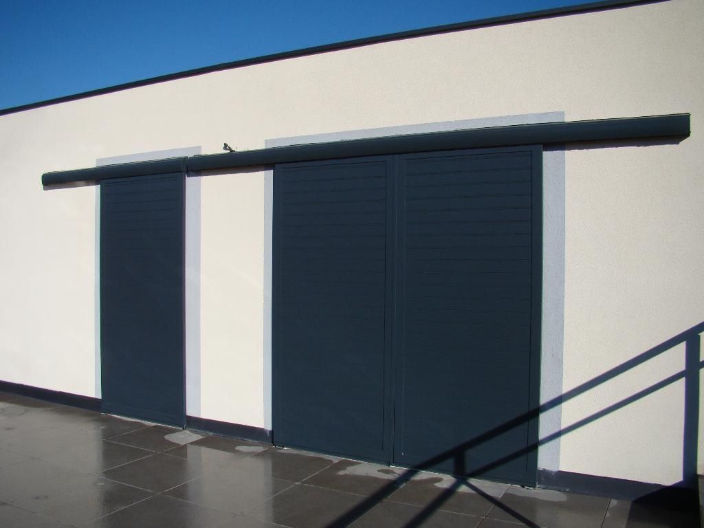 Des portes coulissantes gris foncé, montées sur un rail contre un mur extérieur blanc, donnent sur une terrasse pavée.