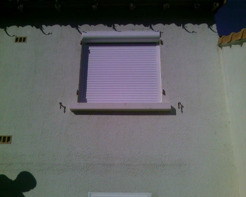 Un mur extérieur blanc d'une maison, avec une fenêtre recouverte d'un volet roulant horizontal blanc fermé.