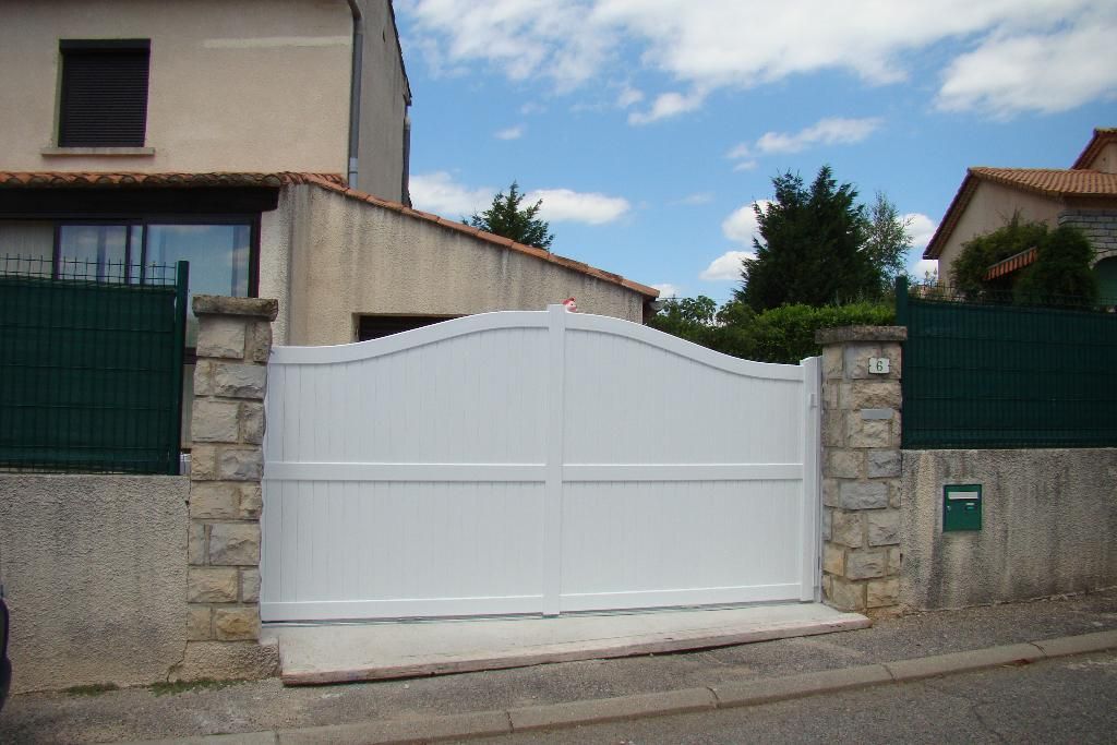 Un portail double blanc en arc de cercle, encadré par deux piliers de pierre, se dresse devant une maison sous un ciel bleu nuageux.