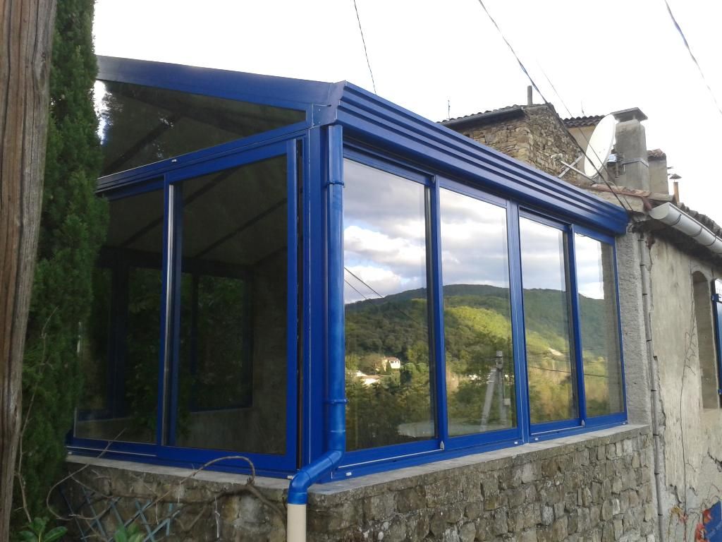 Une véranda aux cadres bleus et aux parois vitrées, attenante à une maison rustique en pierre donnant sur un paysage vallonné.