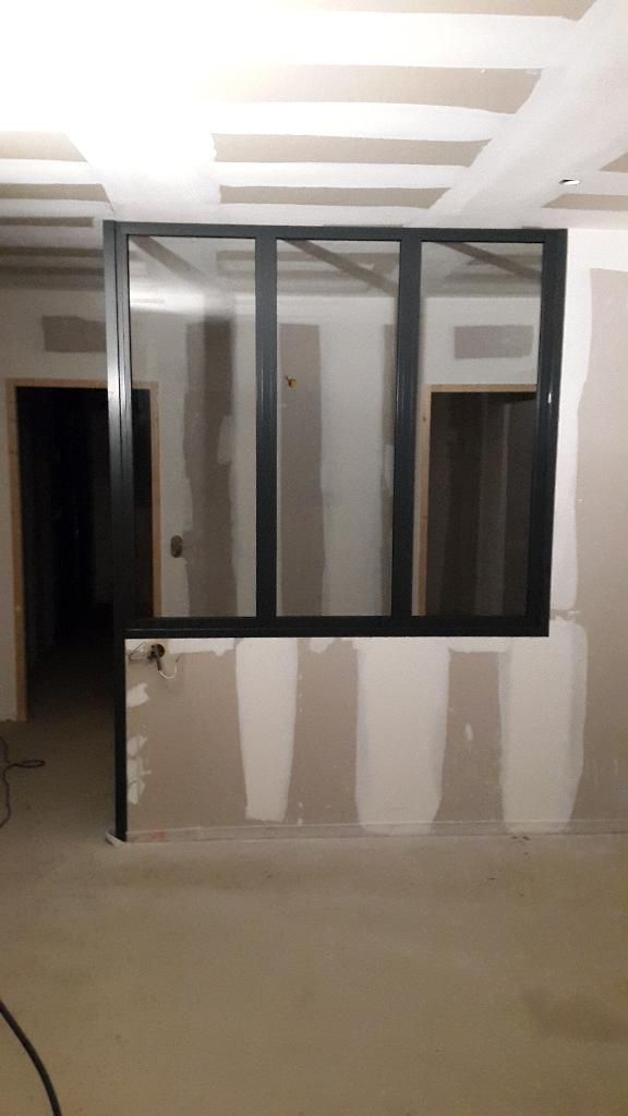 Dans une pièce en construction, une cloison intérieure vitrée à cadre noir repose sur une cloison en plaques de plâtre à mi-hauteur.
