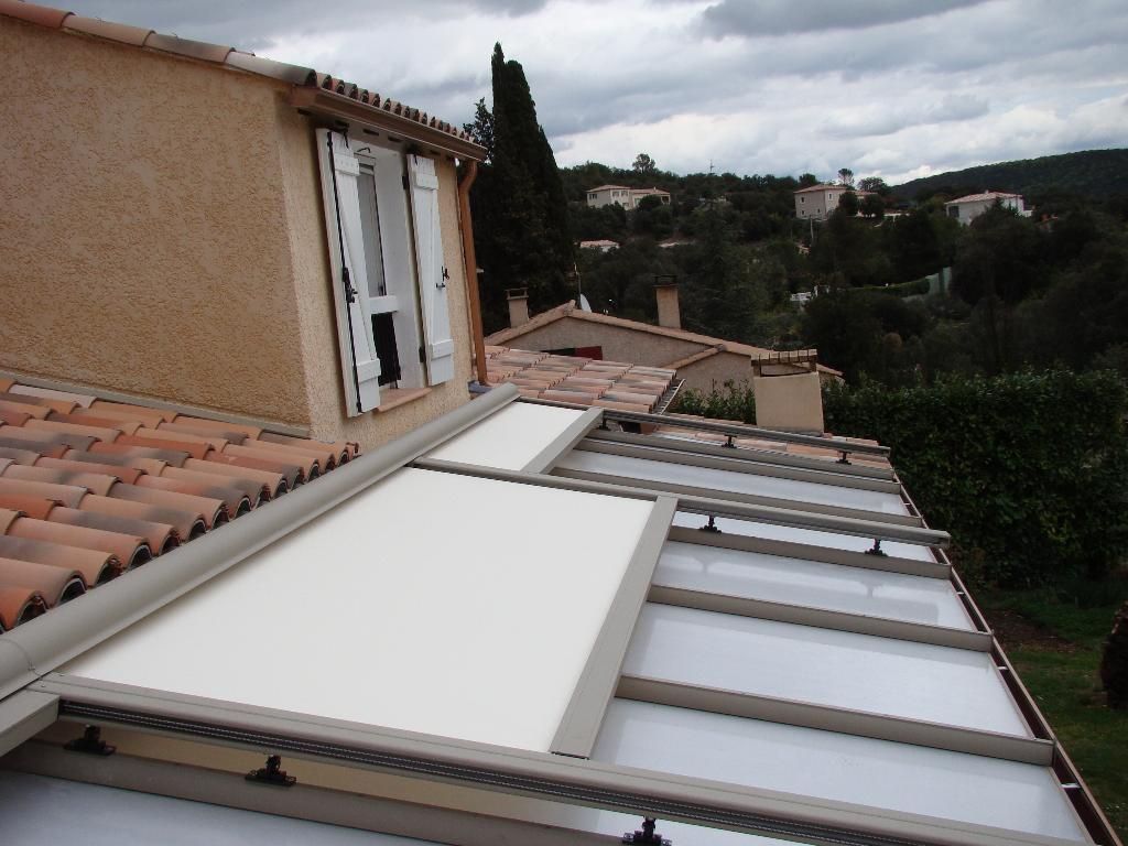 Un store rétractable couleur crème partiellement déployé au-dessus d'une terrasse vitrée jouxtant une maison au toit de tuiles.