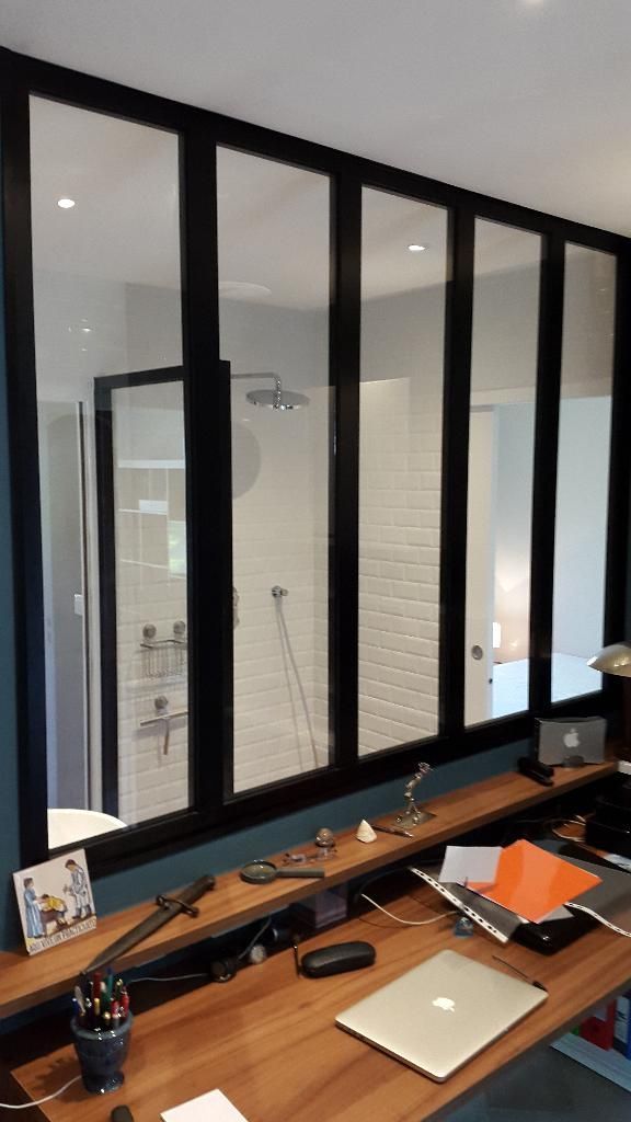 Un espace de travail avec un bureau en bois faisant face à une paroi vitrée à cadre noir, donnant sur l'intérieur carrelé d'une salle de bains.