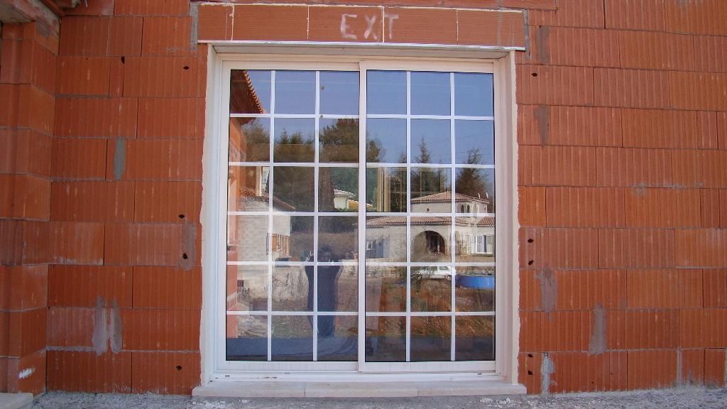 Une porte coulissante en verre à cadre blanc quadrillé, installée dans un mur de briques d'argile rouges creuses en construction.
