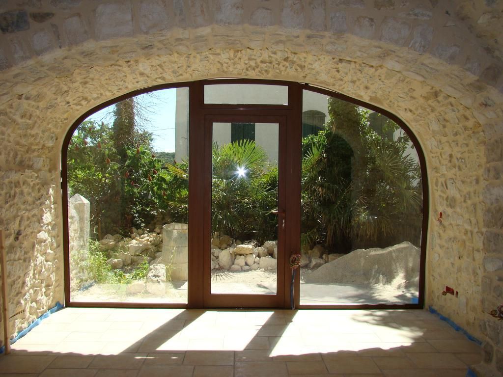 Une porte brune avec une fenêtre rectangulaire, encadrée par une grande arche en pierre, donnant sur un jardin verdoyant.