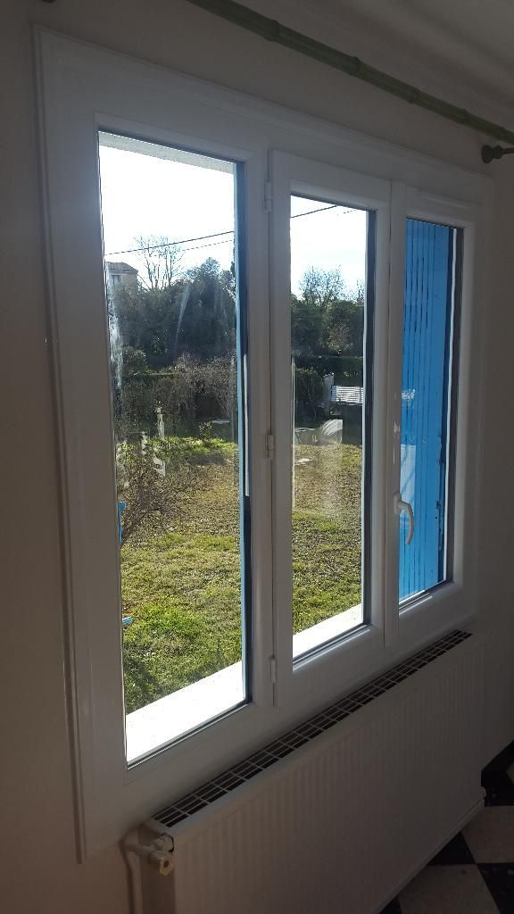 Une fenêtre blanche à trois carreaux donnant sur une cour gazonnée, placée au-dessus d'un radiateur blanc.