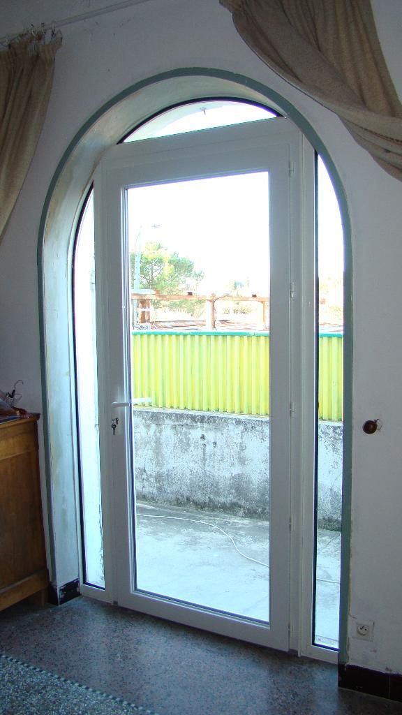 Une porte vitrée cintrée blanche donnant sur un patio extérieur, avec une clôture jaune visible en arrière-plan.