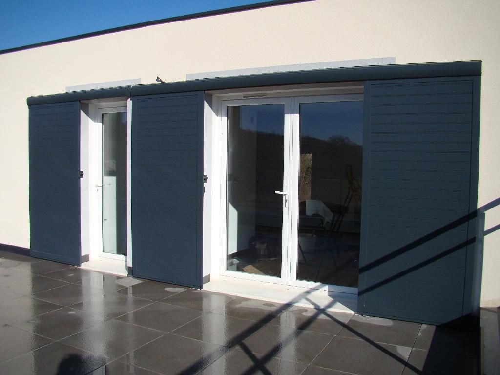 L'extérieur d'un bâtiment moderne présente des portes vitrées blanches et de grands volets coulissants gris foncé sur une terrasse carrelée.