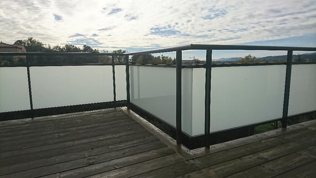 Un coin de terrasse en bois avec une rambarde moderne à cadre noir et panneaux de verre opaque dépoli se détachant sur un ciel nuageux.
