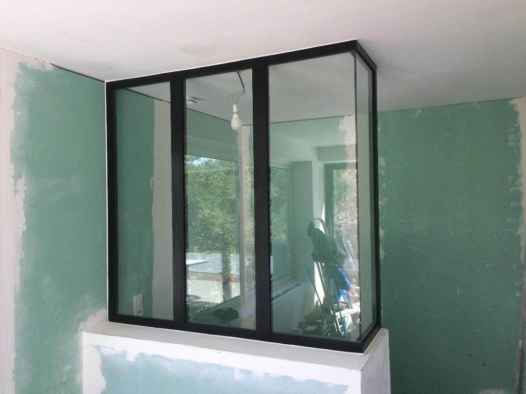 Dans une pièce aux murs vert clair non finis, une cloison vitrée à trois panneaux et à cadre noir repose sur un socle blanc.