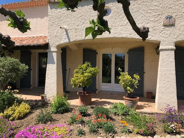 Une maison méditerranéenne ensoleillée avec une façade en stuc crème, des volets bleus, un patio couvert et un jardin de devant luxuriant.