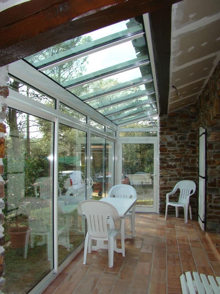 Une véranda avec un toit en verre, un sol en carrelage de terre cuite et des murs en pierre, comprenant une table à manger avec des chaises.