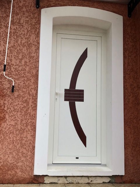Une porte blanche cintrée avec un panneau décoratif central incurvé brun foncé, encastrée dans un mur extérieur texturé brun clair.