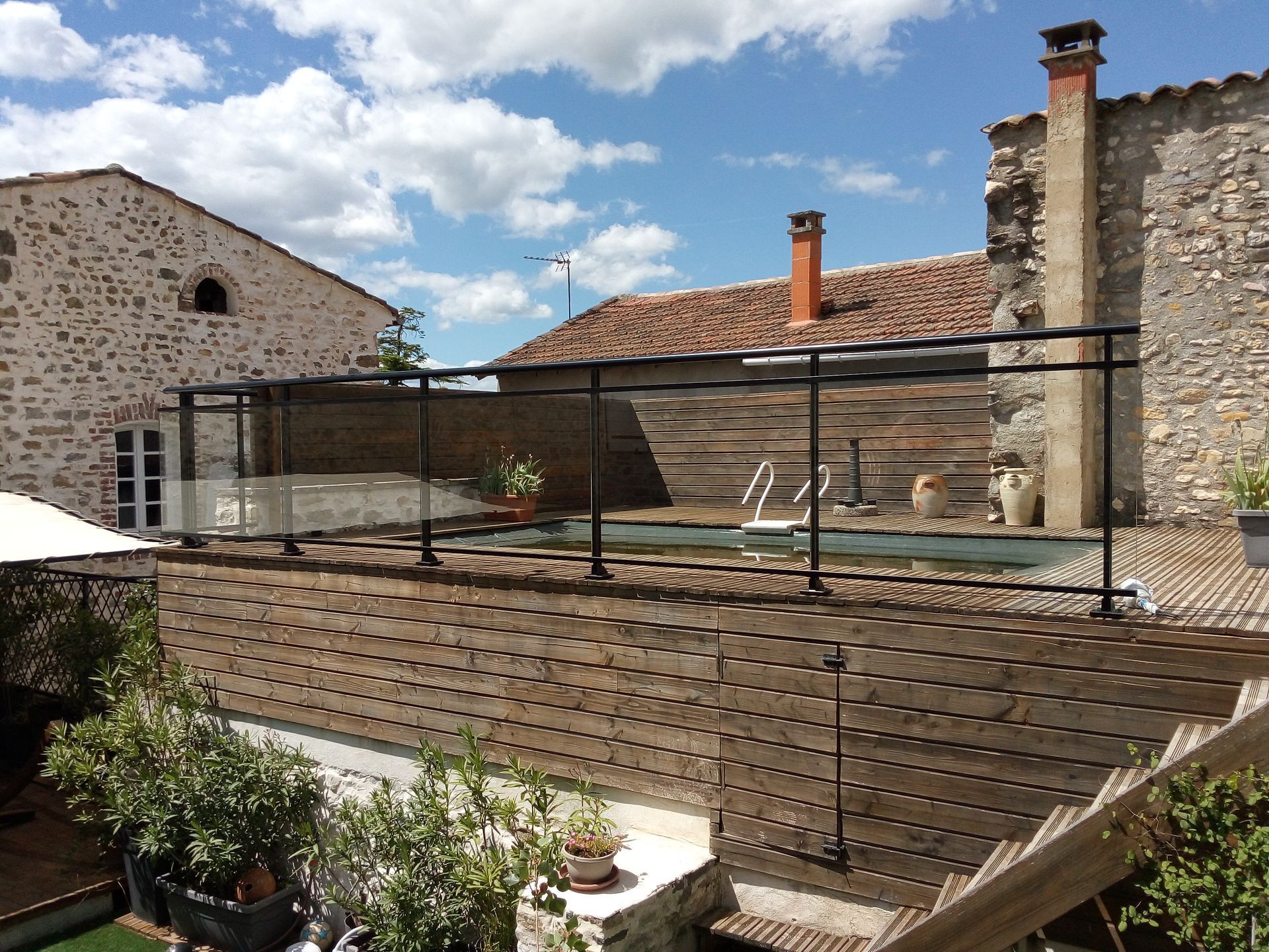 Une terrasse avec piscine, entourée d'une clôture moderne en verre et en métal, située entre des bâtiments rustiques en pierre sous un ciel bleu.
