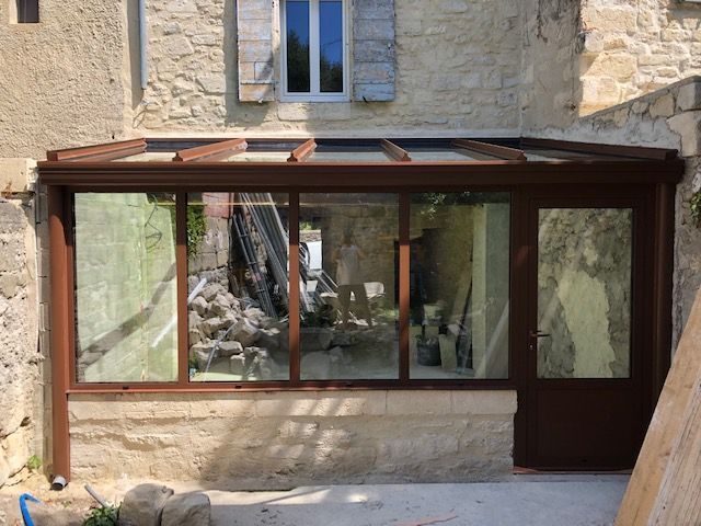 Une véranda avec des cadres bruns et un toit en verre, attenante à un bâtiment en pierre.