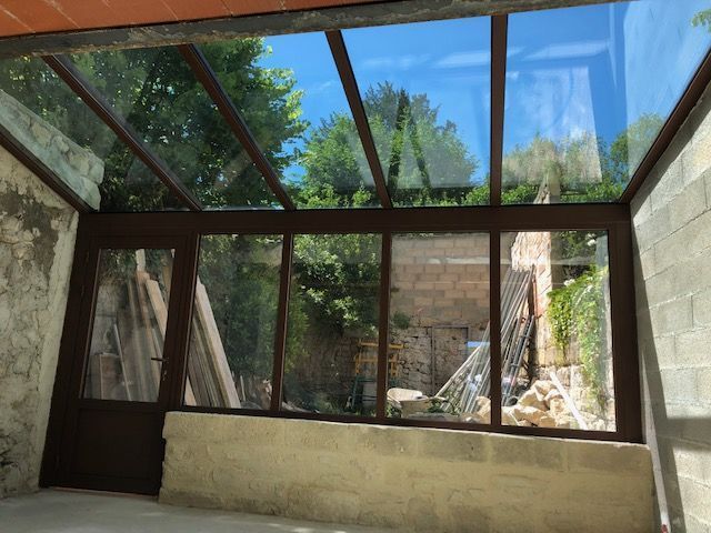 Une véranda vitrée aux cadres métalliques sombres, donnant sur un mur de pierre et des arbres verdoyants dans un jardin.