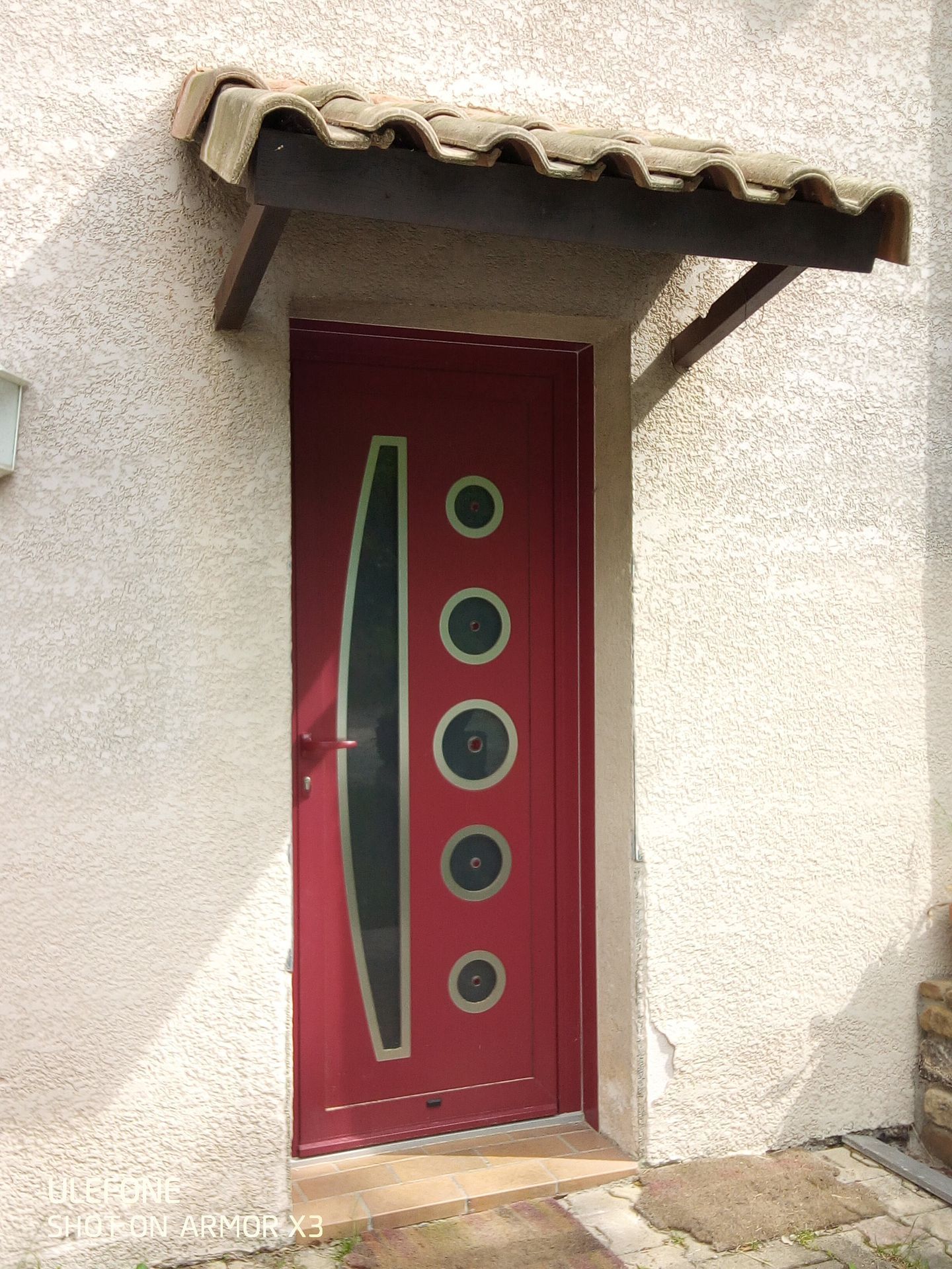 Une porte d'entrée rouge moderne avec un panneau de verre vertical et quatre fenêtres circulaires, sous un petit auvent carrelé de brun.