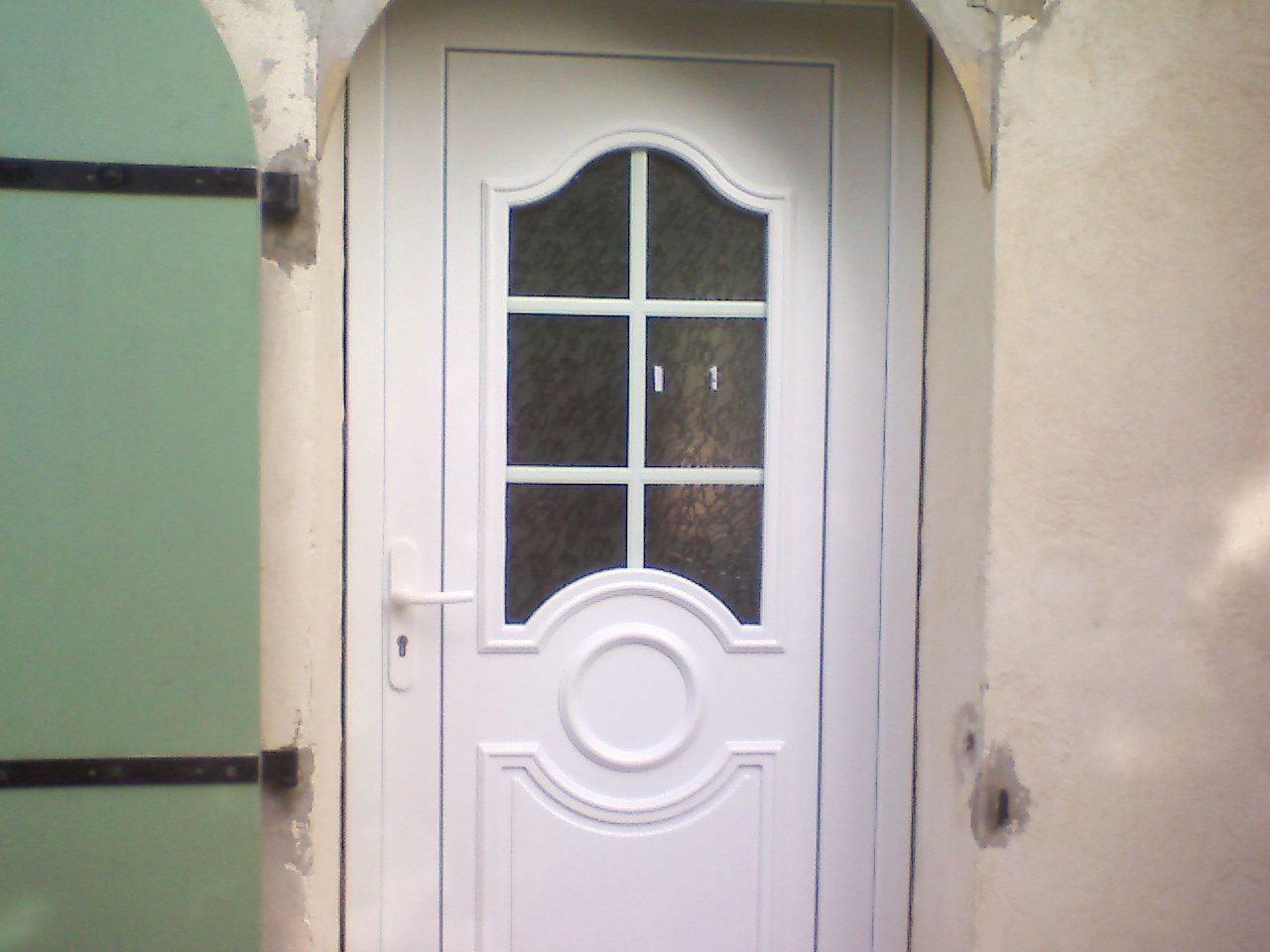 Une porte blanche avec une fenêtre à six carreaux en partie supérieure, un panneau central décoratif circulaire et une poignée latérale dans une simple niche d'entrée.