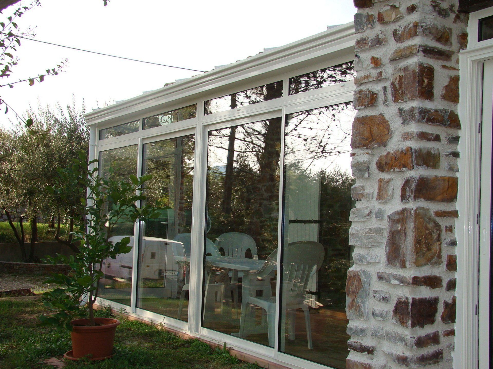 Une véranda vitrée à ossature blanche, adossée à un bâtiment en pierre et donnant sur un jardin.