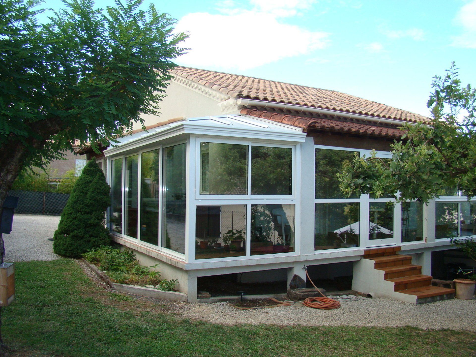 Une maison avec une grande véranda vitrée à cadre blanc, entourée d'arbres et d'une cour gravillonnée.