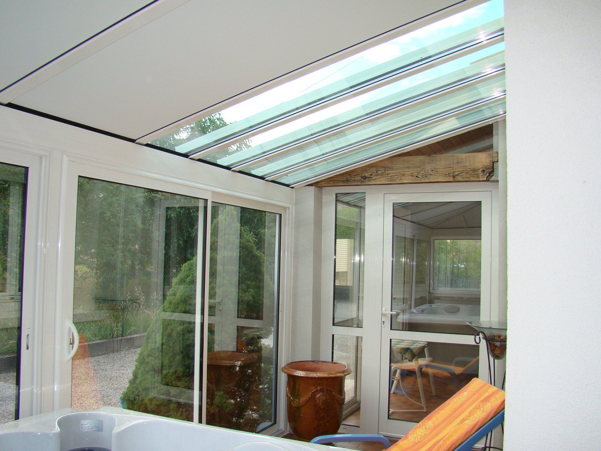 Véranda avec toit en verre, portes coulissantes, grand pot en céramique et vue sur une pièce secondaire avec jacuzzi.