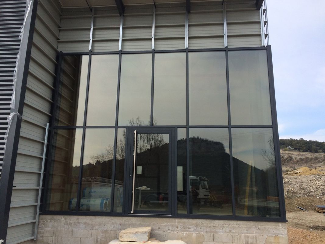 Une grande vitrine en verre avec un cadre en métal noir, installée sur un socle en pierre, à côté d'un revêtement métallique sur la façade d'un bâtiment.