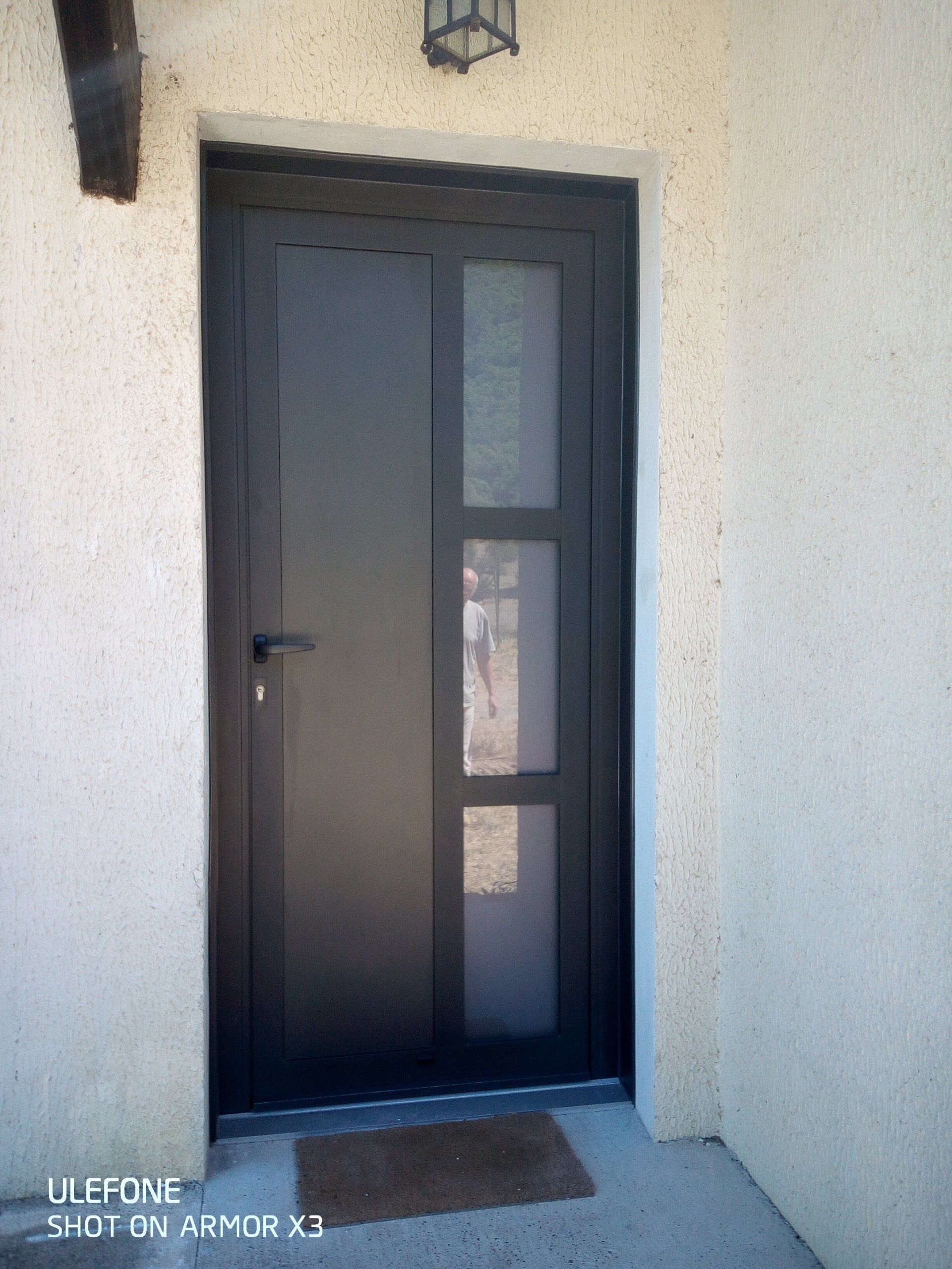 Une porte d'entrée moderne à cadre sombre, composée de trois panneaux de verre verticaux encastrés dans un mur extérieur en stuc beige.