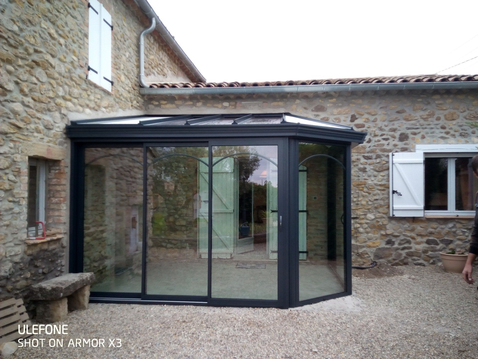 Une véranda moderne en verre à cadre sombre, attenante à une maison rustique en pierre avec des volets blancs, sur une terrasse en gravier.