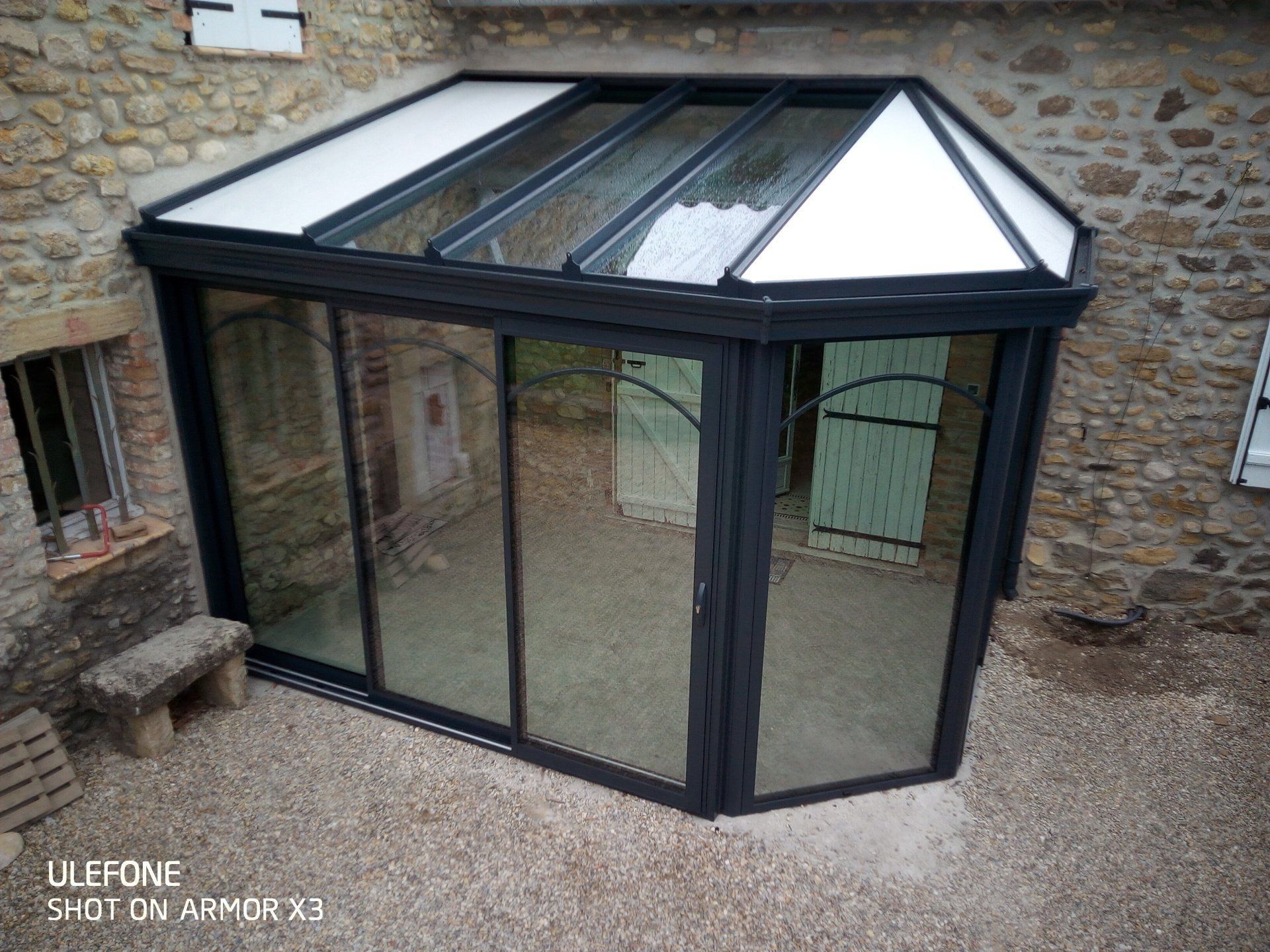 Une véranda à ossature sombre, adossée à un mur en pierre, dotée de grandes portes coulissantes en verre et d'un toit en verre à plusieurs panneaux.