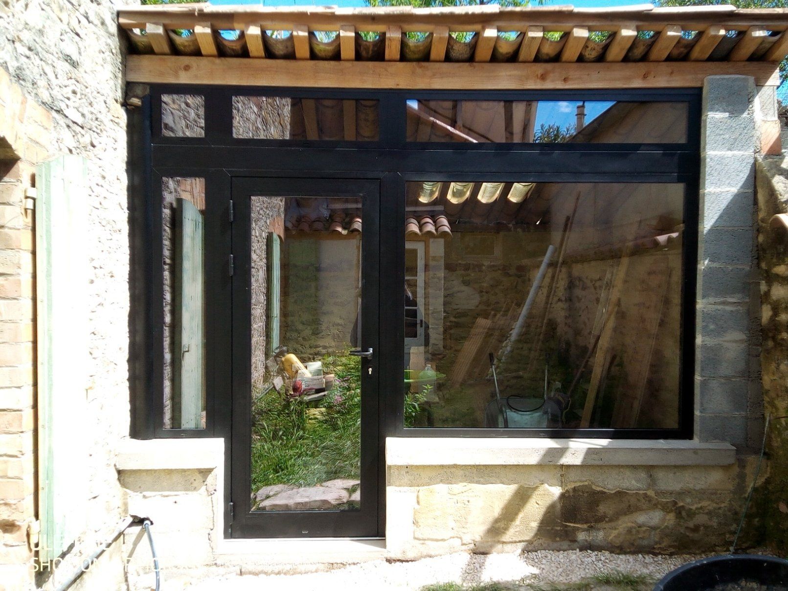 Une porte et une fenêtre vitrées à cadre noir, encastrées dans un mur de pierre sous un porche au toit en bois.