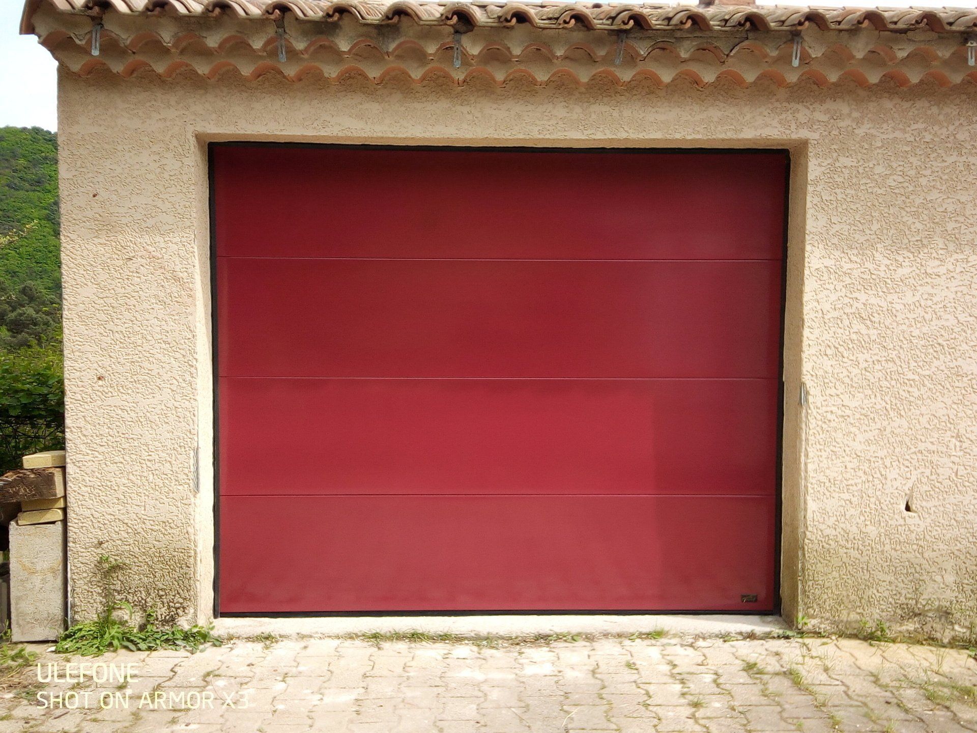 Une porte de garage rectangulaire fermée, composée de quatre panneaux horizontaux d'un rouge profond, encastrée dans un mur texturé beige clair.