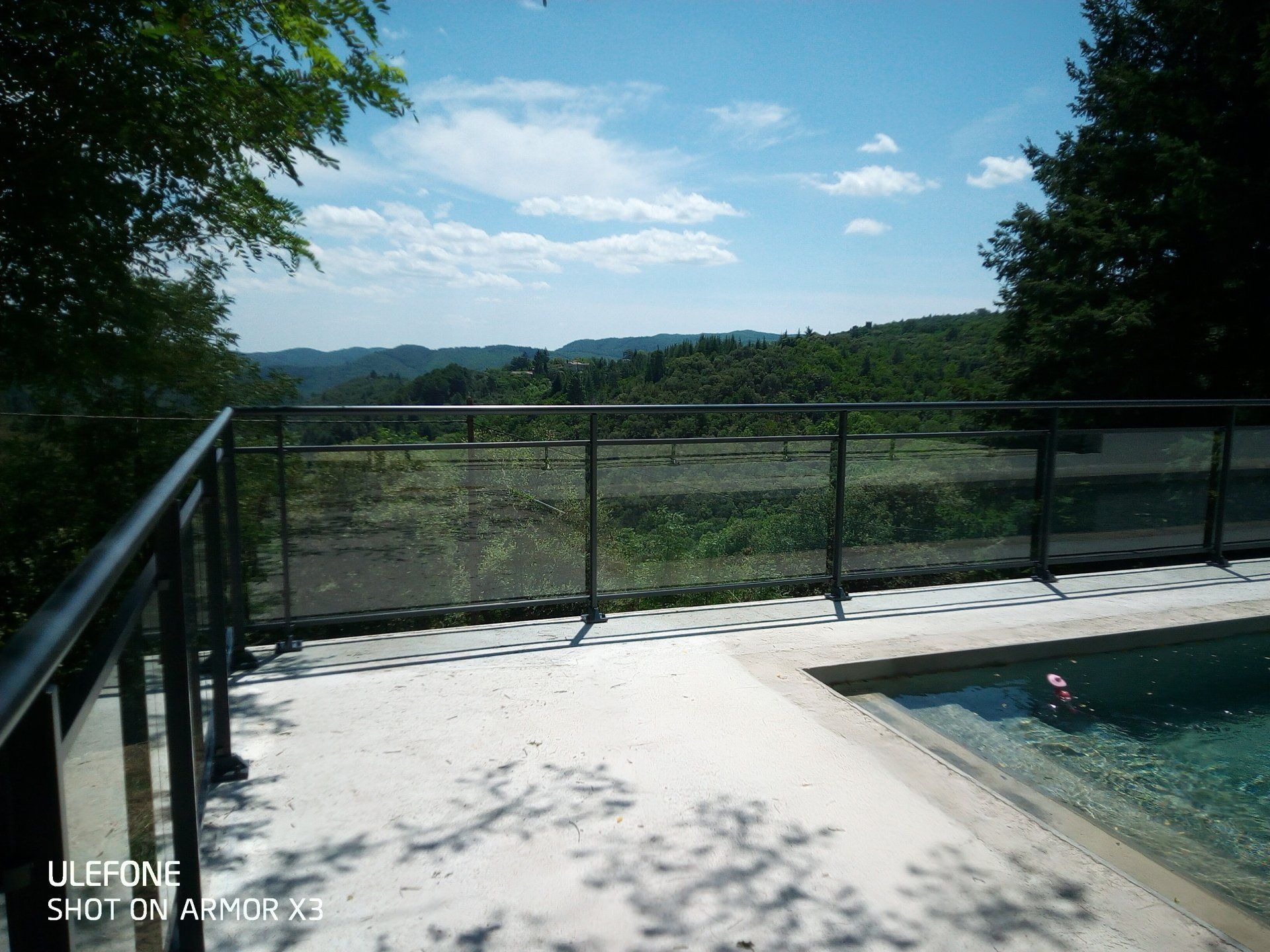 Une terrasse de piscine avec une rambarde en métal donnant sur une vallée verdoyante sous un ciel d'un bleu éclatant.