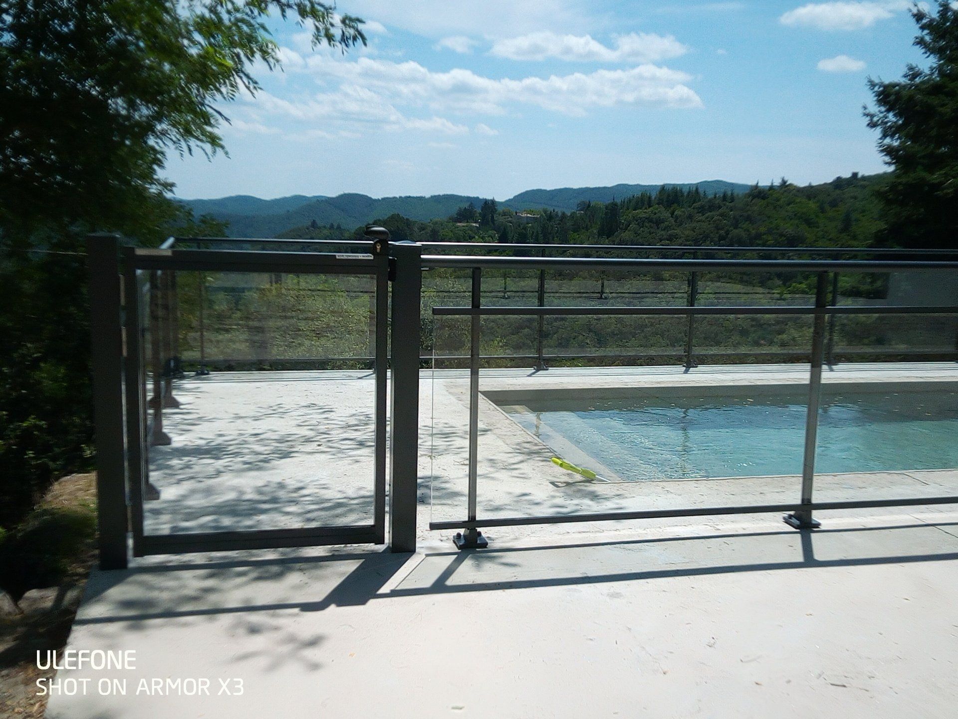 Une clôture métallique entoure une terrasse de piscine en béton donnant sur un paysage pittoresque de collines verdoyantes sous un ciel bleu.