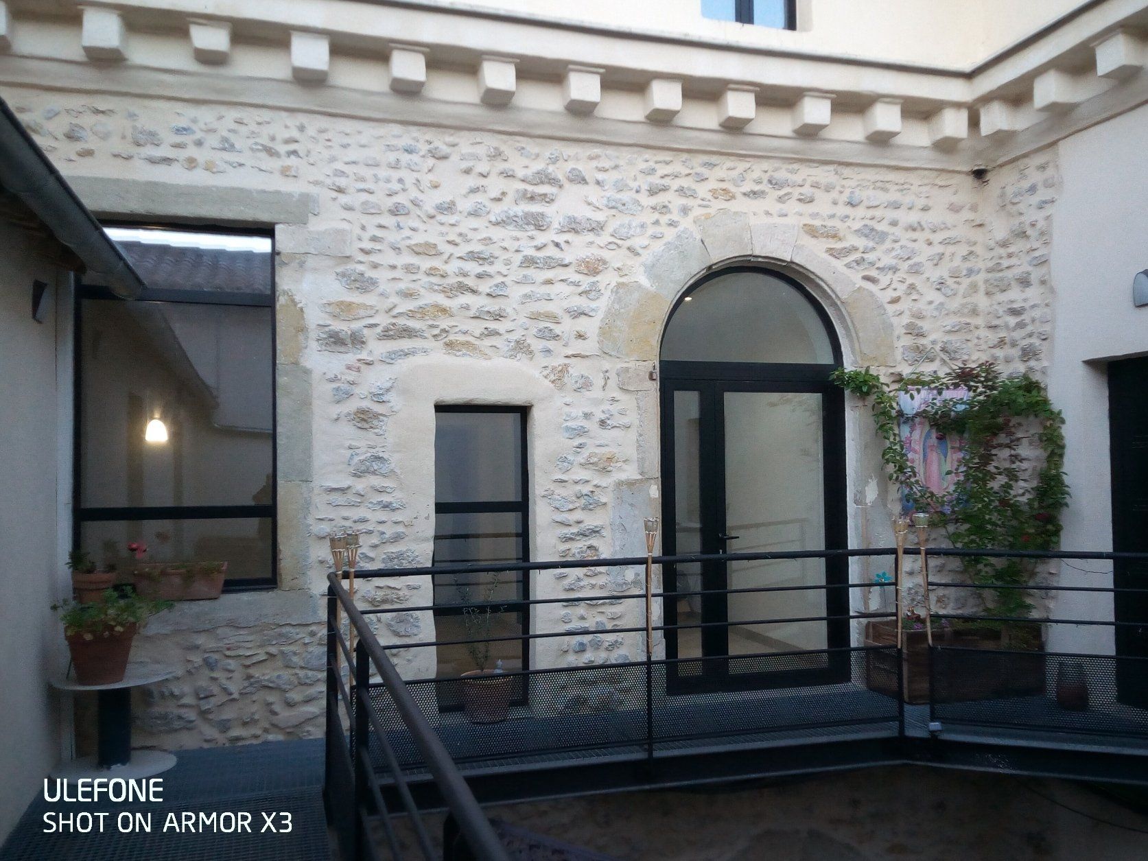 Une cour en pierre agrémentée d'un balcon en métal noir, d'une haute porte vitrée cintrée, d'une fenêtre plus petite et de plantes en pot.