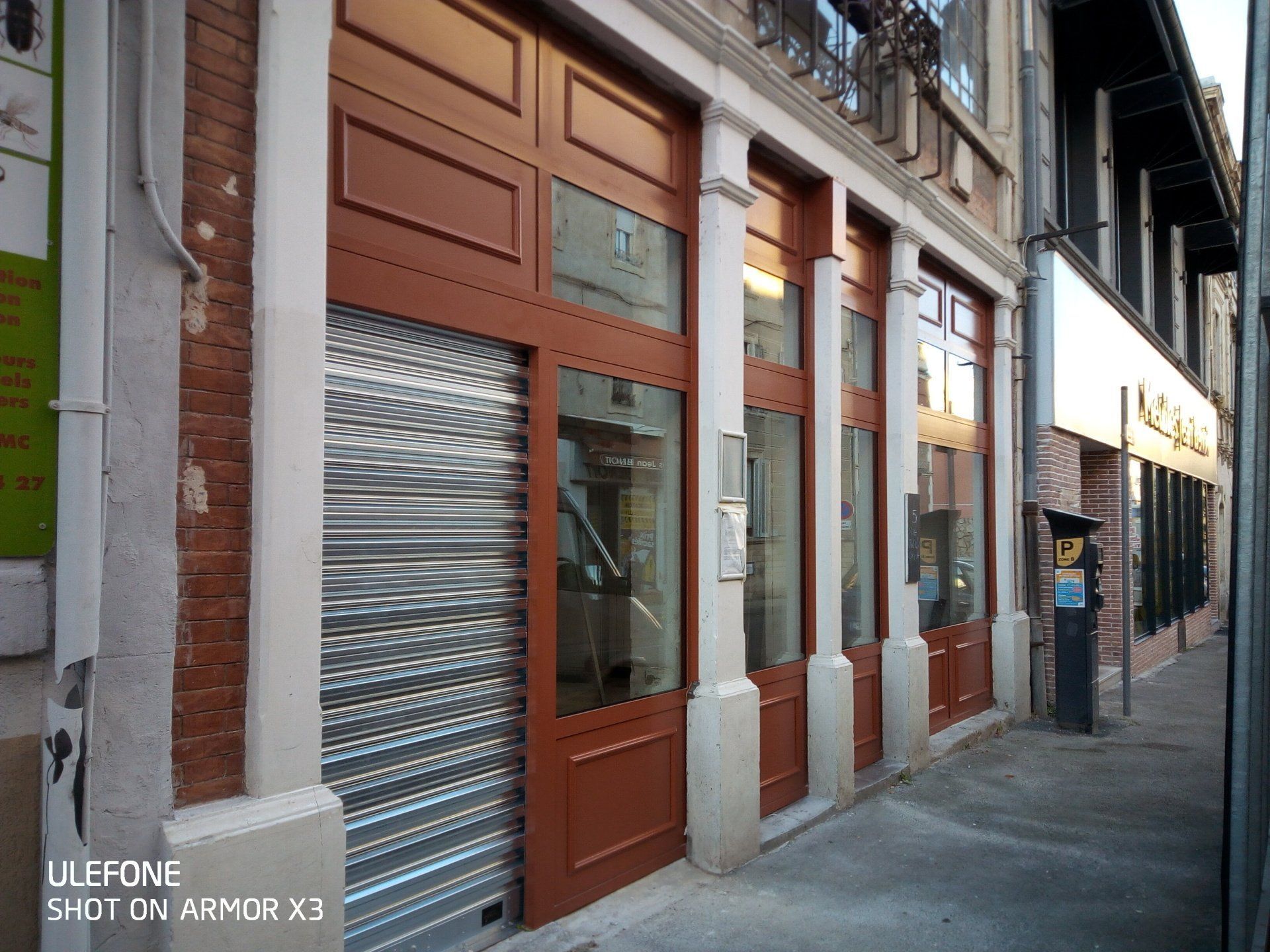 Une façade de magasin avec des boiseries rouge-brun, des panneaux de verre et une porte roulante métallique, située sur un trottoir en béton.